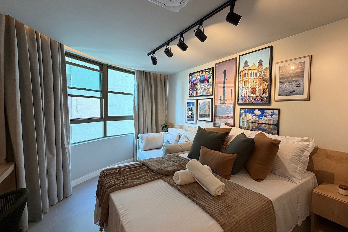 A hotel room with a bed, a bedside table, a lamp, a window with curtains, and wall art.