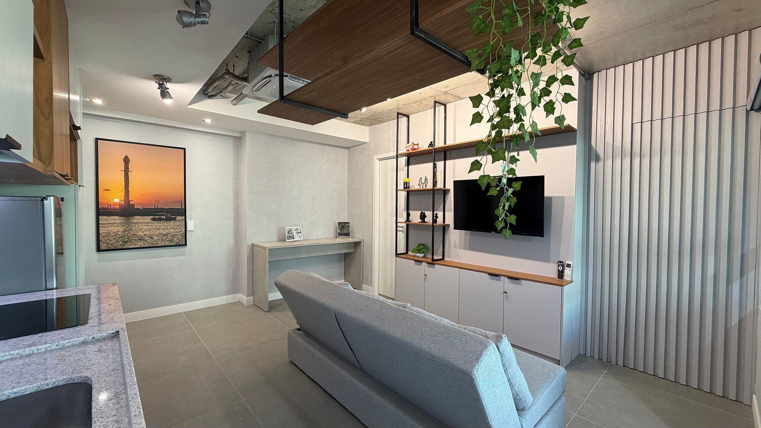 A modern living room with a grey sofa, a flat-screen TV mounted on a wall, and a wooden shelf with decorative items.