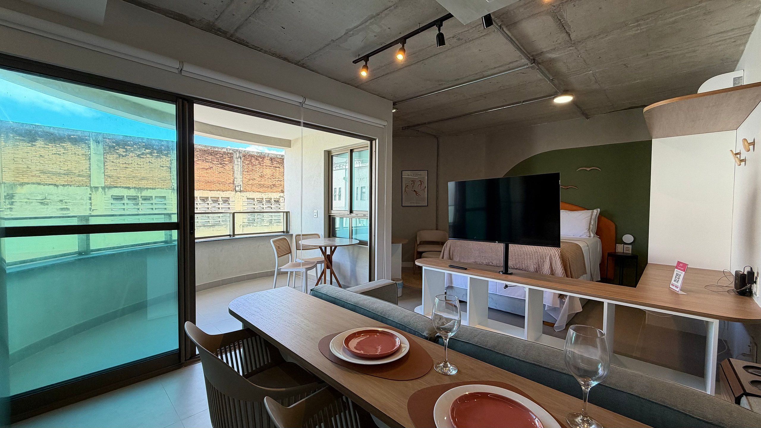 Habitación de hotel con balcón, zona de comedor con sillas y mesa, cama con televisor y vistas a la ciudad.
