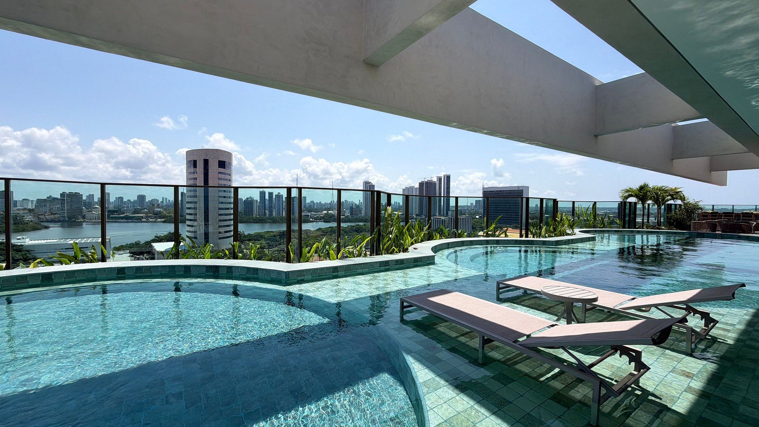 A rooftop infinity pool with lounge chairs, surrounded by a glass railing, overlooking a city skyline and a body of water.