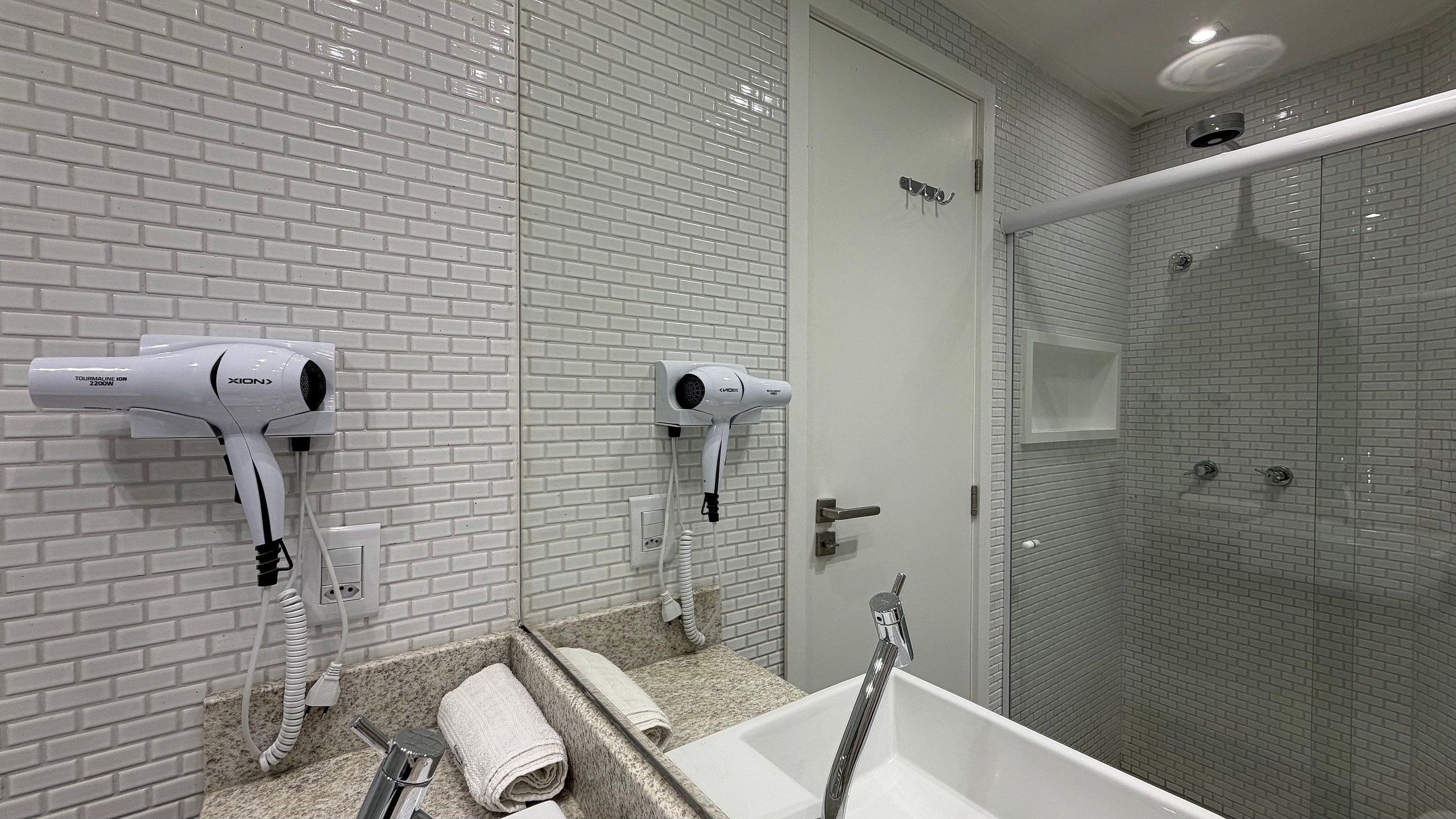A bathroom with two hair dryers mounted on the wall, a sink with a mirror, and a glass shower enclosure.