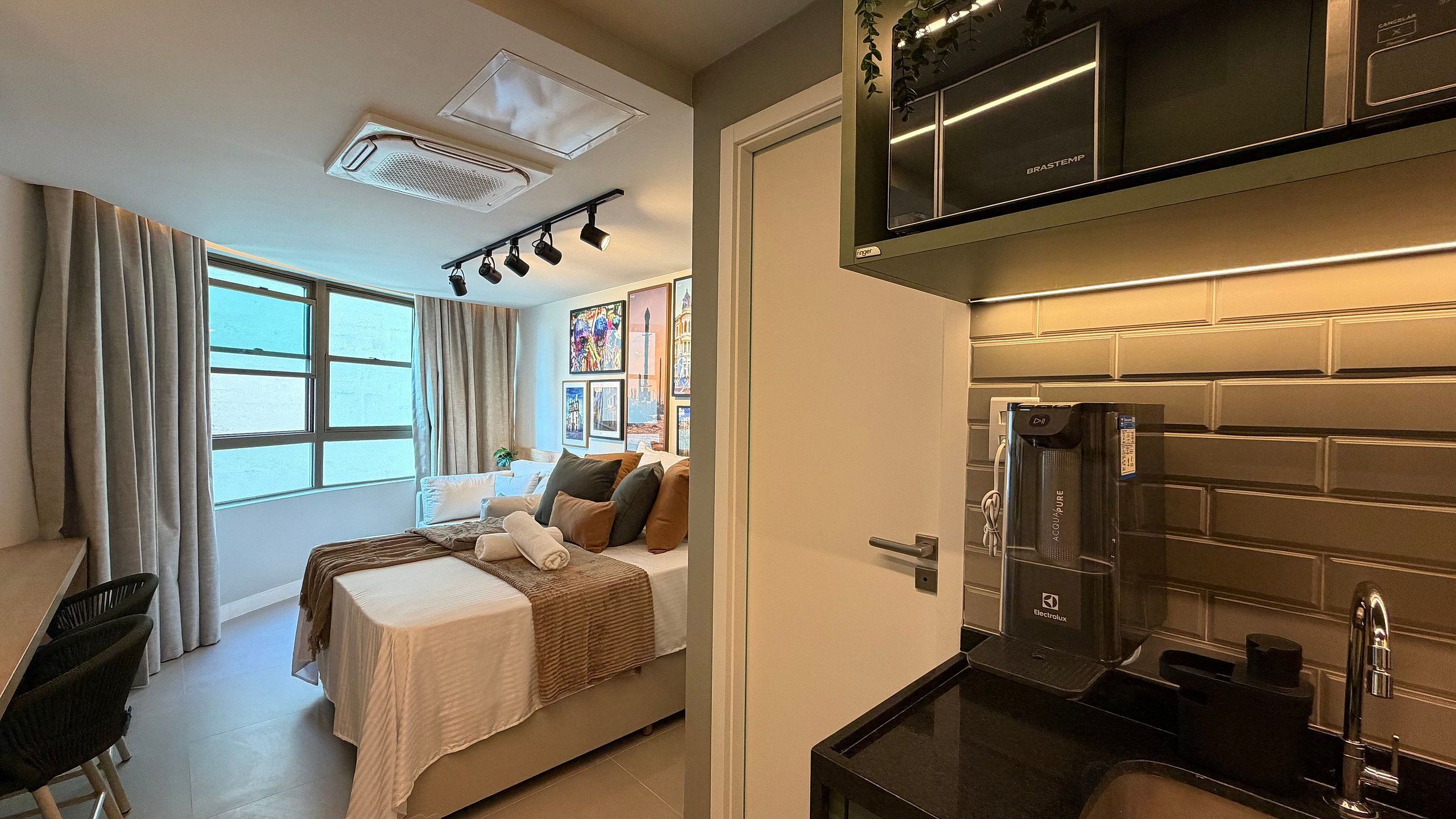 A modern hotel room with a bed, a desk, a chair, a window with curtains, a kitchenette with a refrigerator, and a ceiling light fixture.