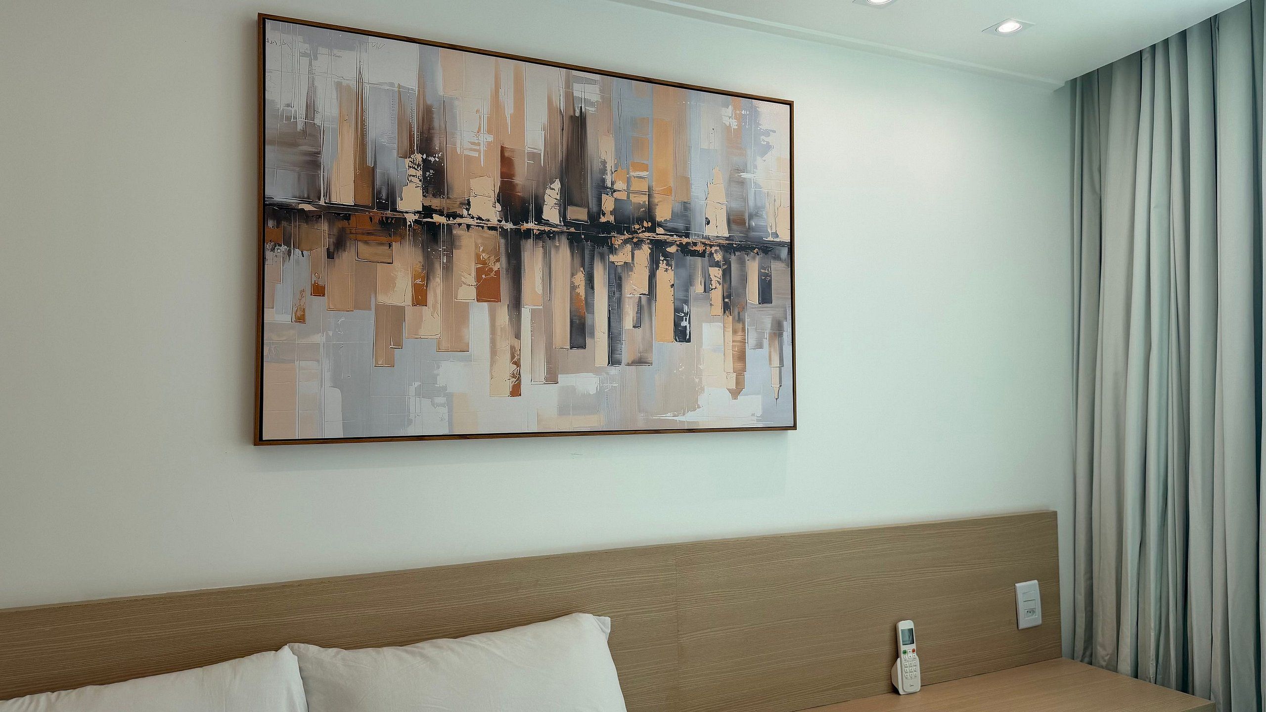 A hotel room with a large abstract painting on the wall, a wooden headboard, a white pillow, and a bedside table with a remote control.