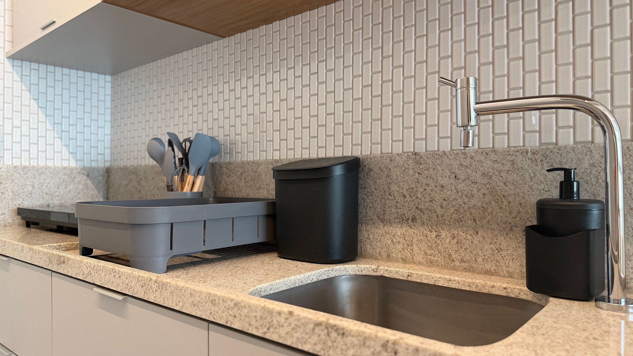 A kitchen with a granite countertop, a stainless steel sink, a black trash bin, a soap dispenser, and a utensil holder.