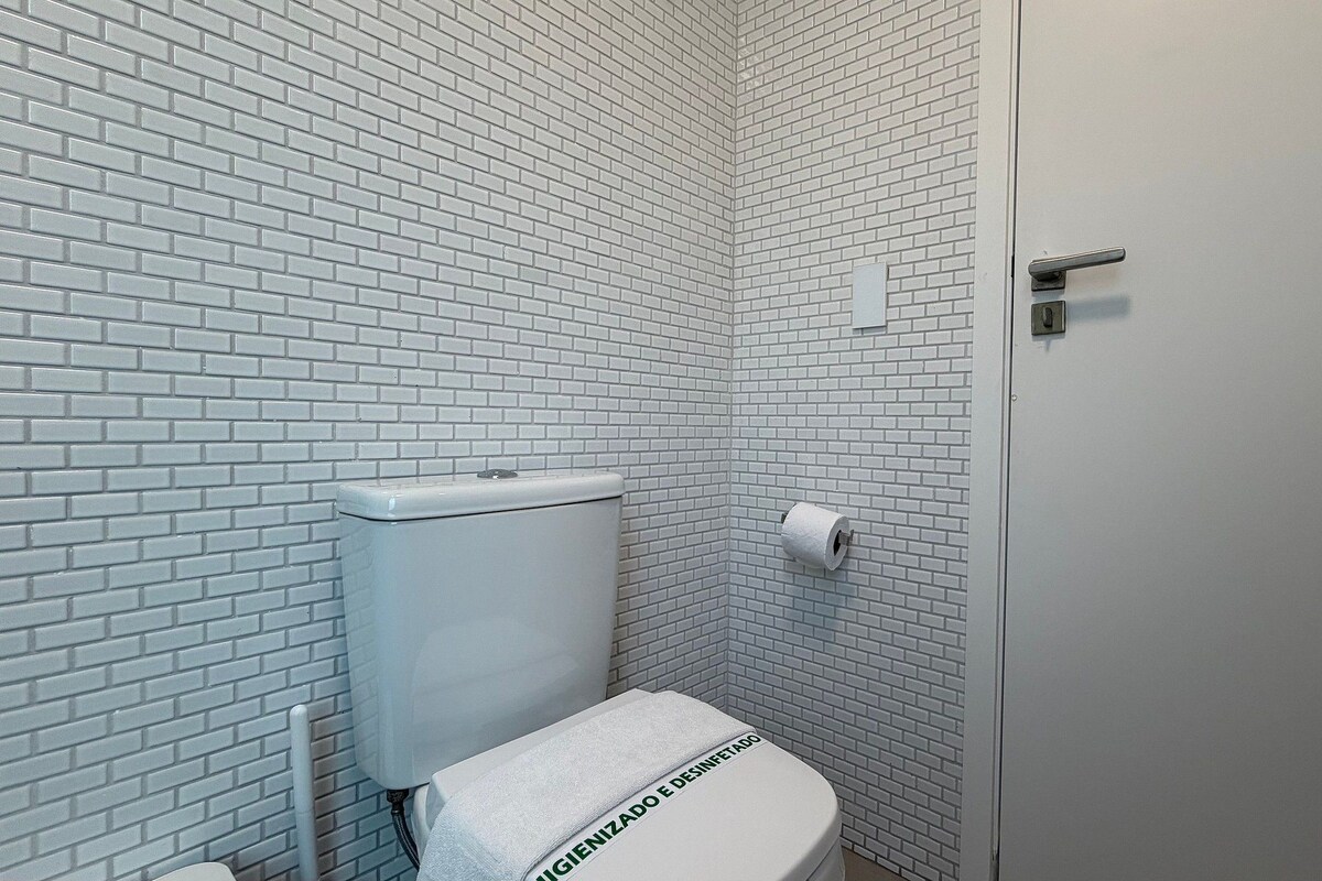 A small, white-tiled bathroom with a toilet, a roll of toilet paper, and a door.