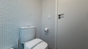 A small, white-tiled bathroom with a toilet, a roll of toilet paper, and a door.