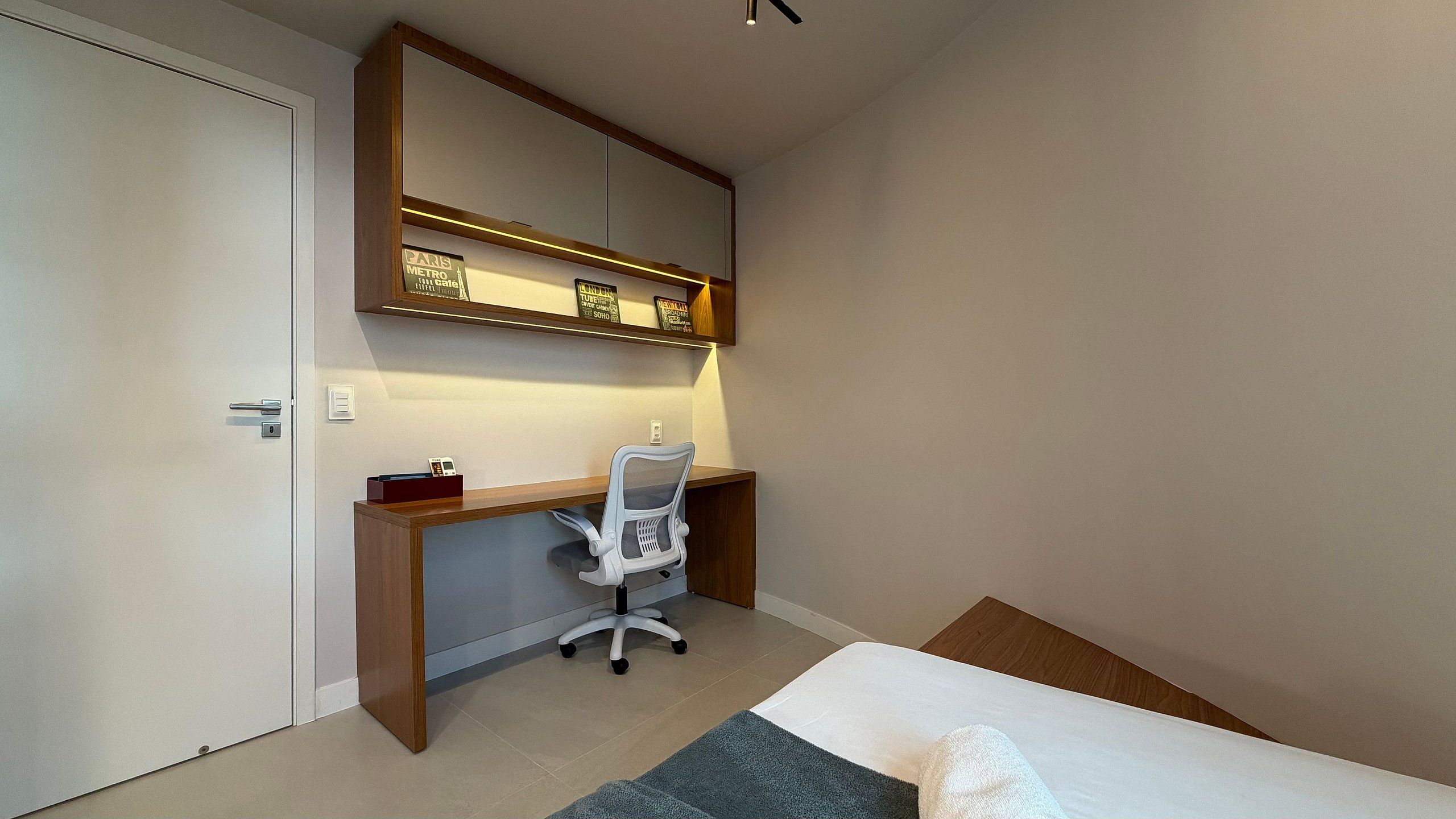 Ein Schlafzimmer mit einem Bett, einem Schreibtisch und einem Stuhl. Eine Tür und ein Regal mit Gegenständen sind ebenfalls vorhanden.