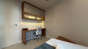 A bedroom with a bed, a desk, and a chair. There is a door and a shelf with items on it.