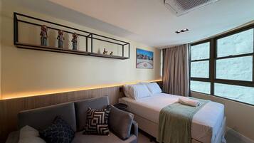 Habitación de hotel con cama, sofá, estante adosado a la pared con objetos decorativos, un cuadro y una ventana con cortinas.
