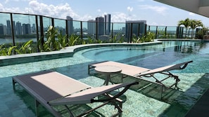 A rooftop infinity pool with lounge chairs and a table, surrounded by greenery and a city skyline view.