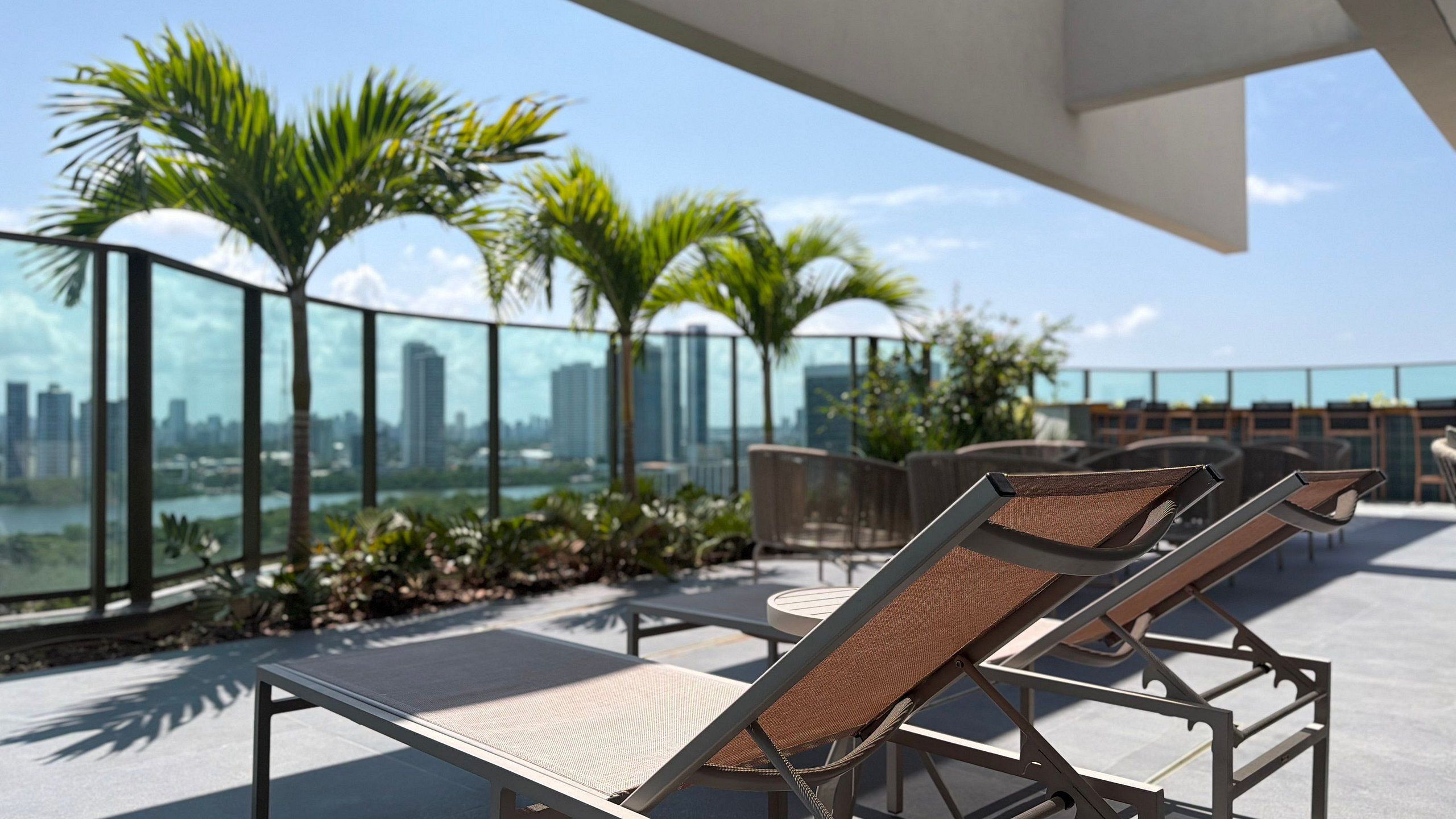 A rooftop terrace with lounge chairs, a glass balustrade, and a cityscape view.