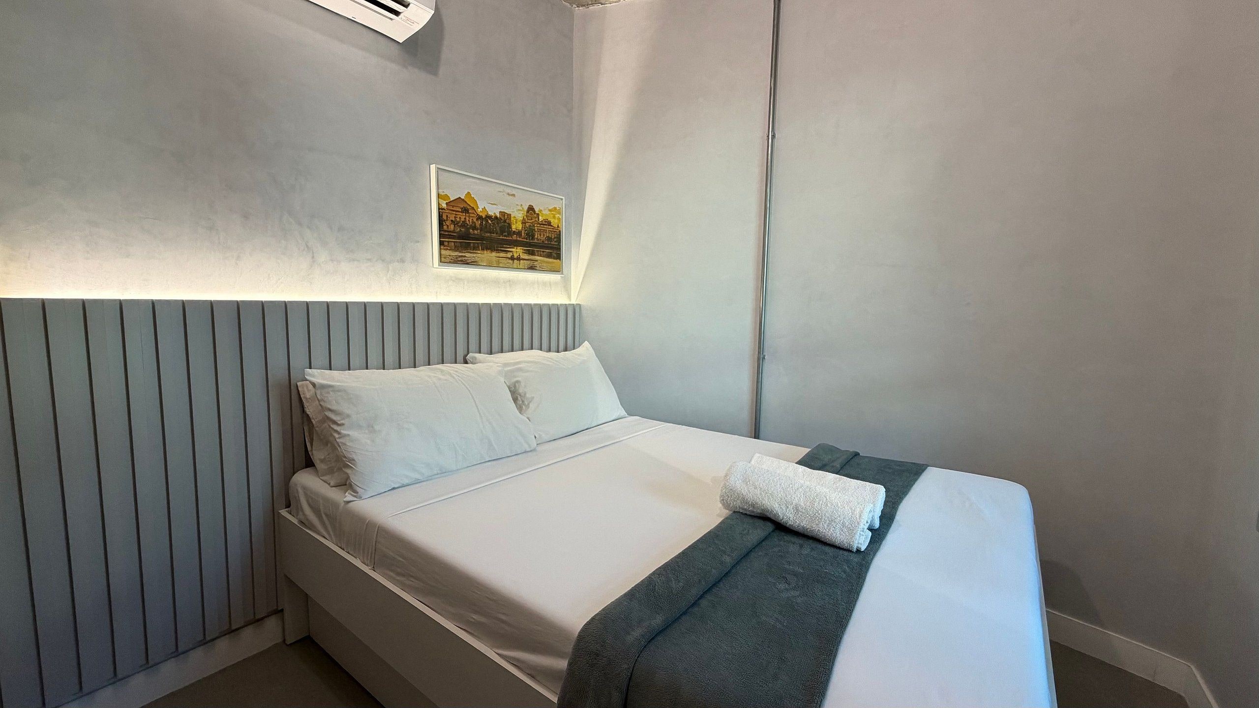 Una cama individual con ropa de cama blanca, cabecera gris y un cuadro en la pared.