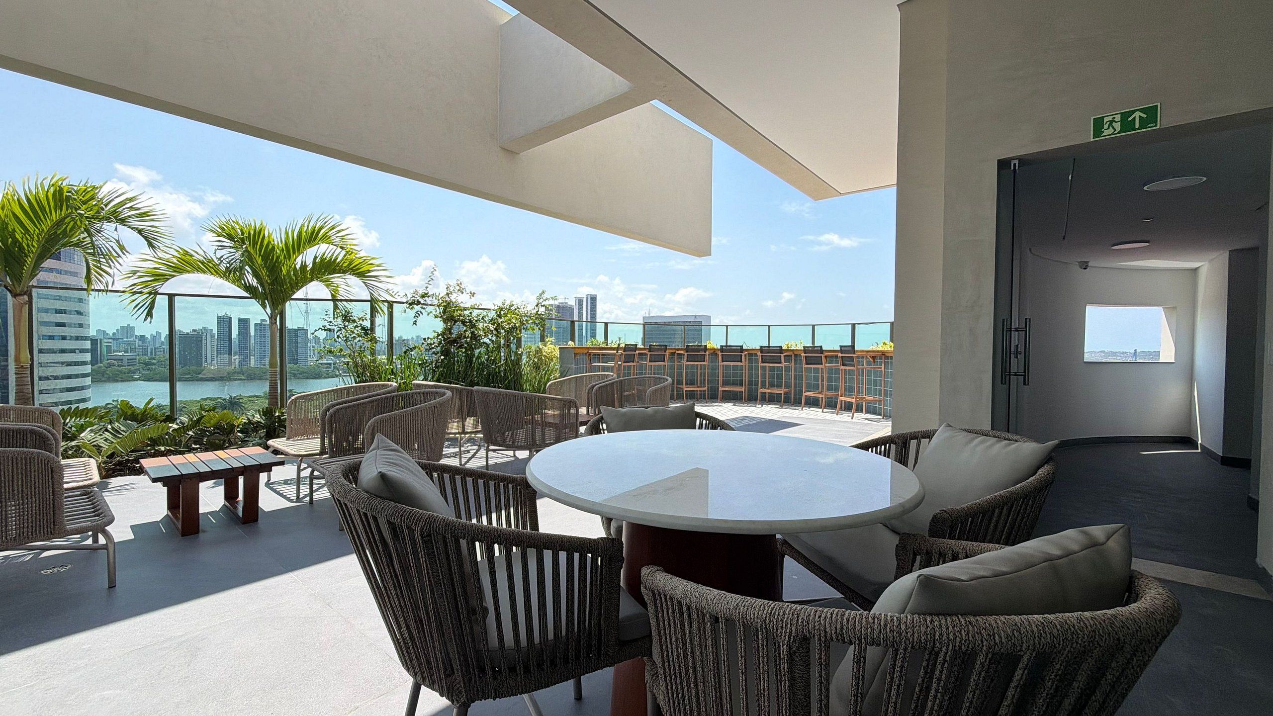 A rooftop lounge with wicker furniture, a round table, and a view of the city skyline.