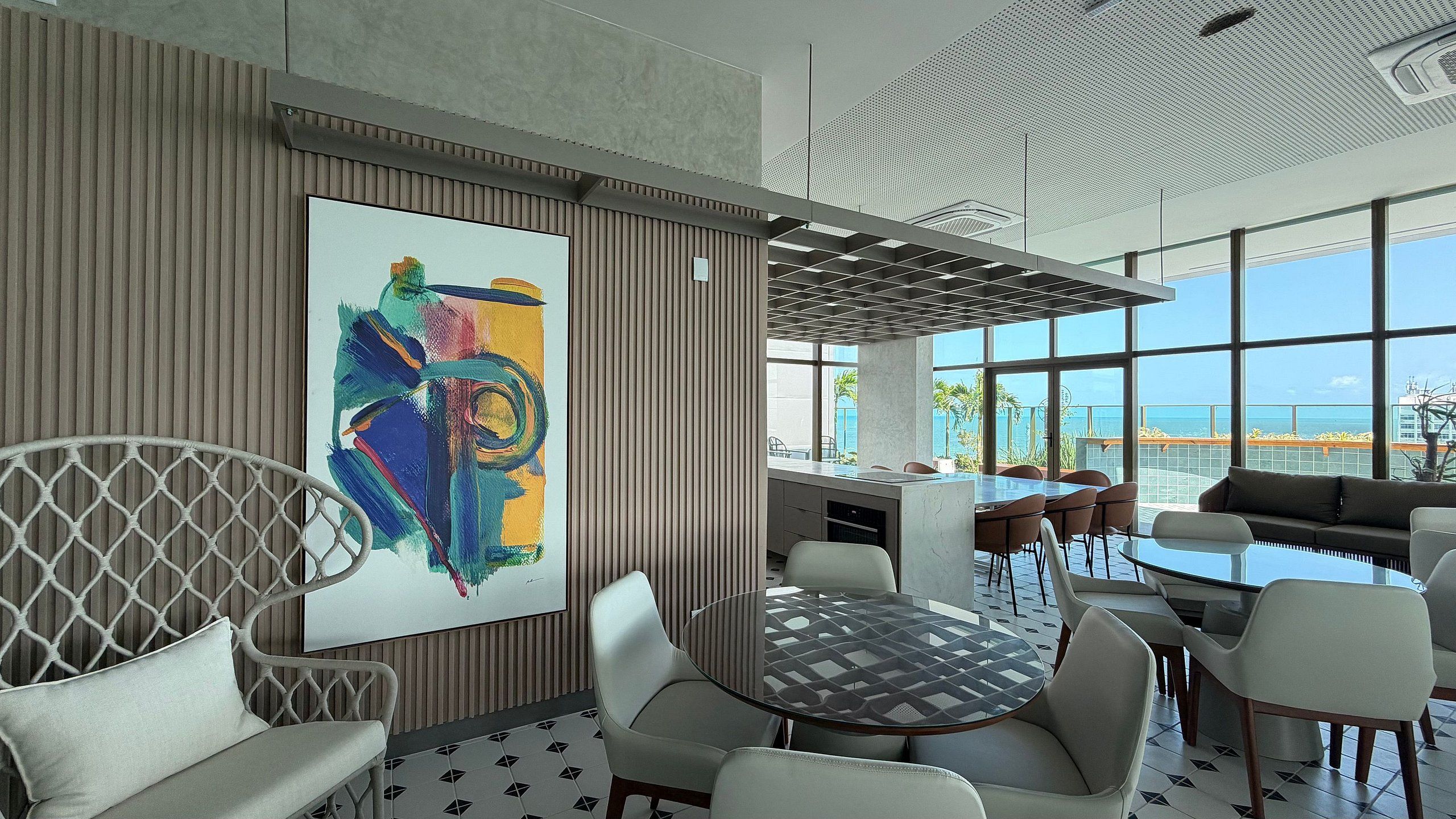 Un interior moderno con un cuadro abstracto grande, un área de comedor con mesas redondas y vistas al océano a través de ventanas amplias.