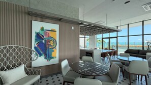 Un interior moderno con un cuadro abstracto grande, un área de comedor con mesas redondas y vistas al océano a través de ventanas amplias.