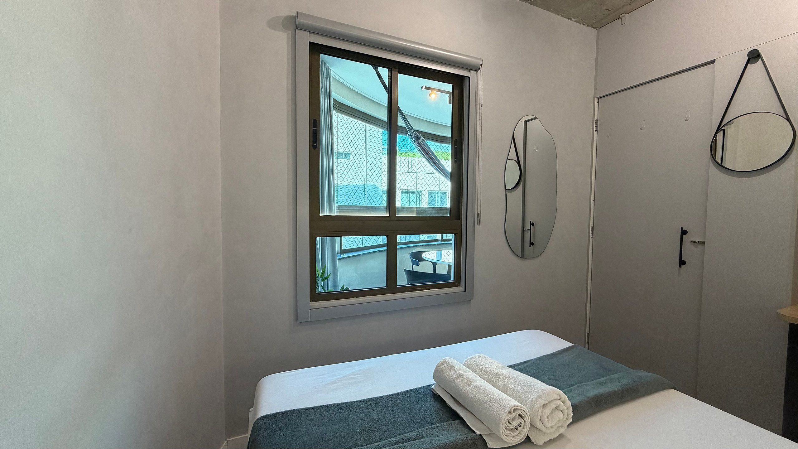 Una cama individual con colcha blanca y aguamarina, una ventana con vista a un edificio y una puerta con toallero.