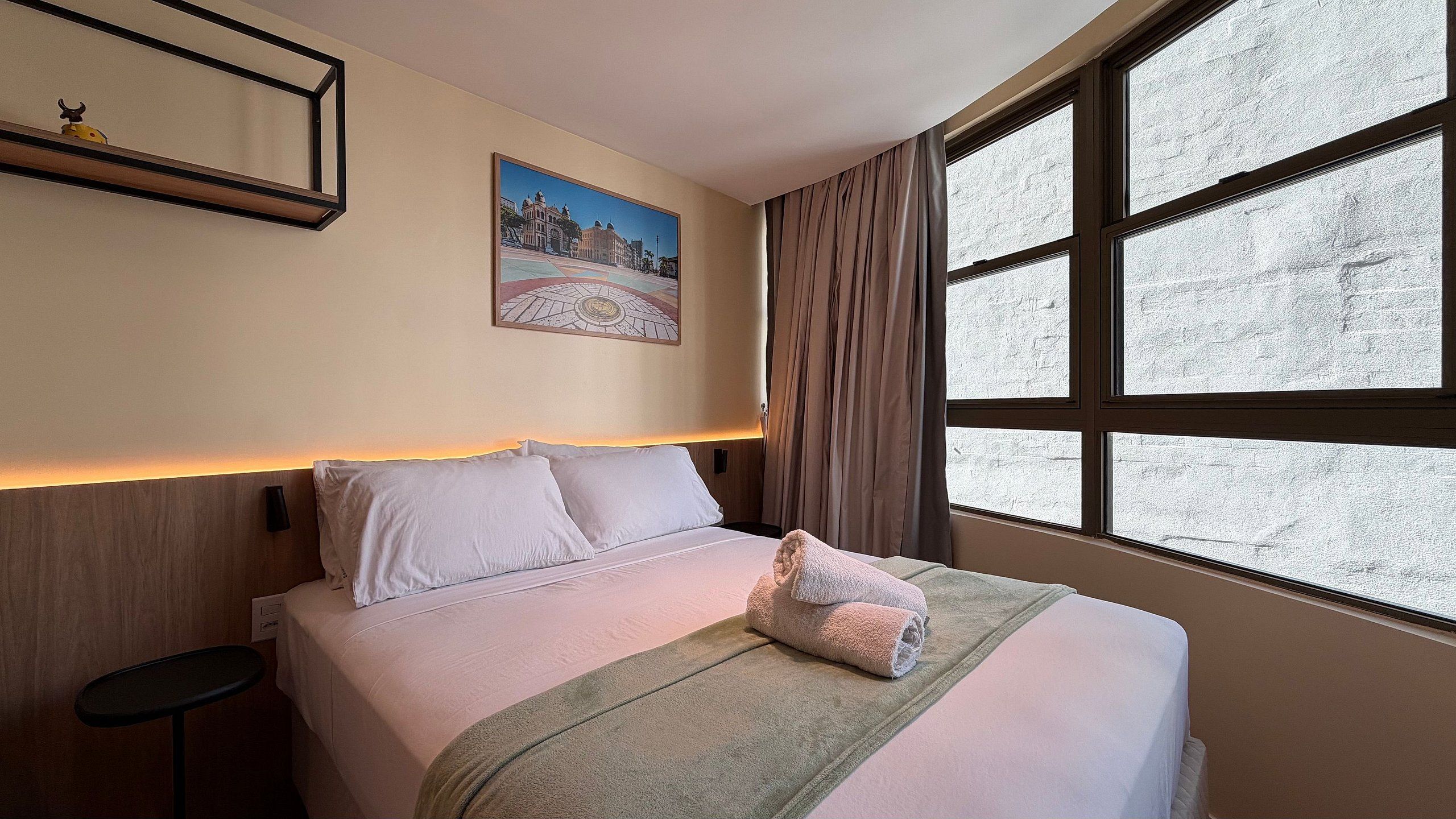 A hotel room with a bed, a nightstand, a window with curtains, and a wall-mounted picture.