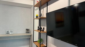 Un salon moderne avec un grand téléviseur à écran plat fixé au mur, des étagères en bois ornées d’objets décoratifs et un tableau encadré placé sur une étagère inférieure.