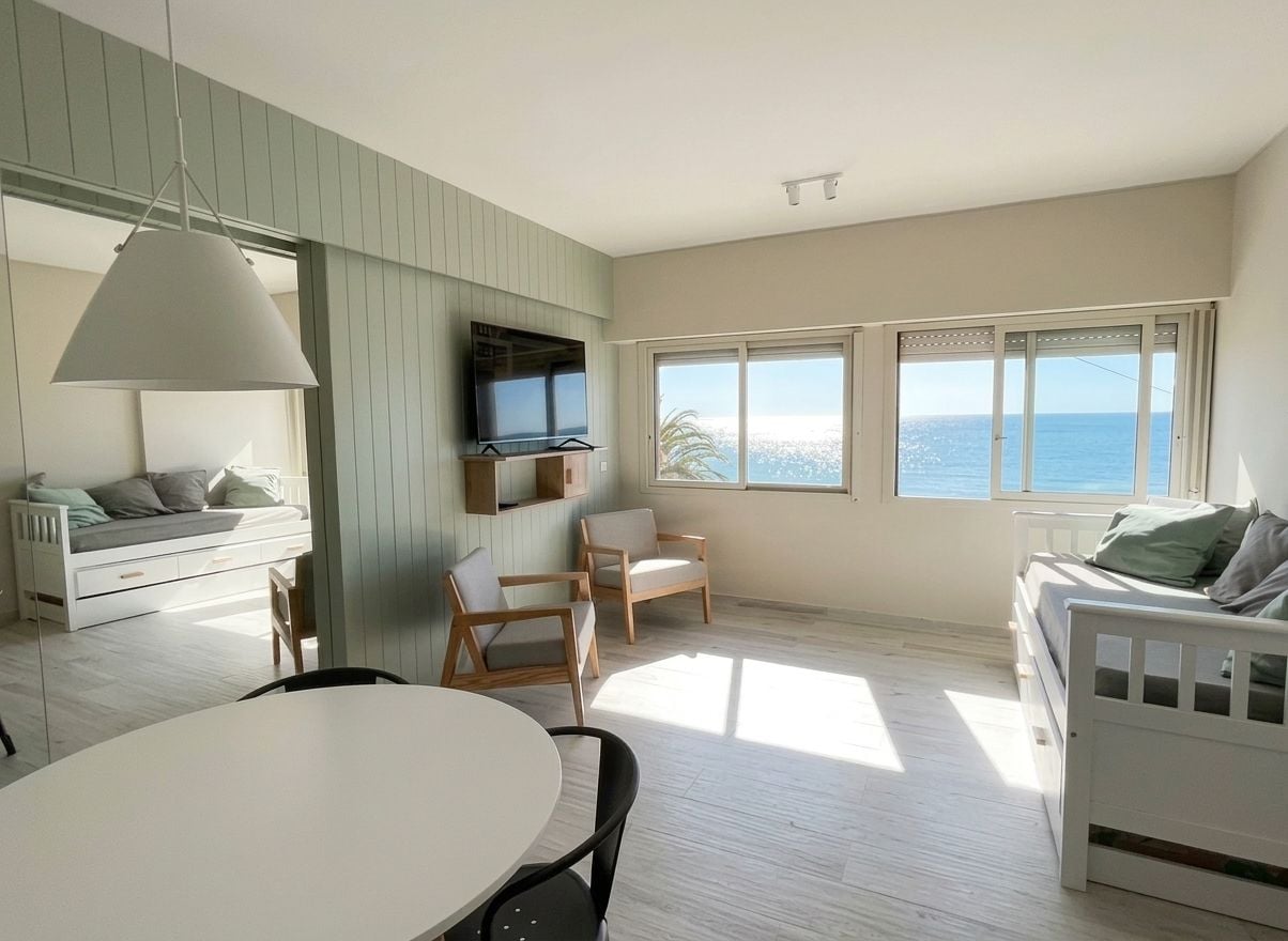 A modern living room with a dining area, a white sofa, a TV mounted on the wall, and large windows with a view of the sea.