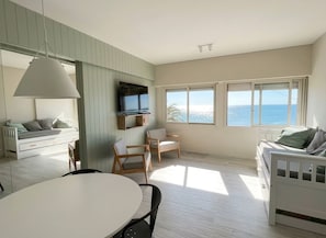 A modern living room with a dining area, a white sofa, a TV mounted on the wall, and large windows with a view of the sea.