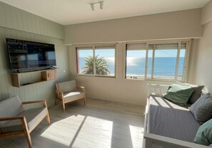 Ein Zimmer mit einem großen Fernseher, einer Couch, zwei Sesseln und Meerblick durch große Fenster.