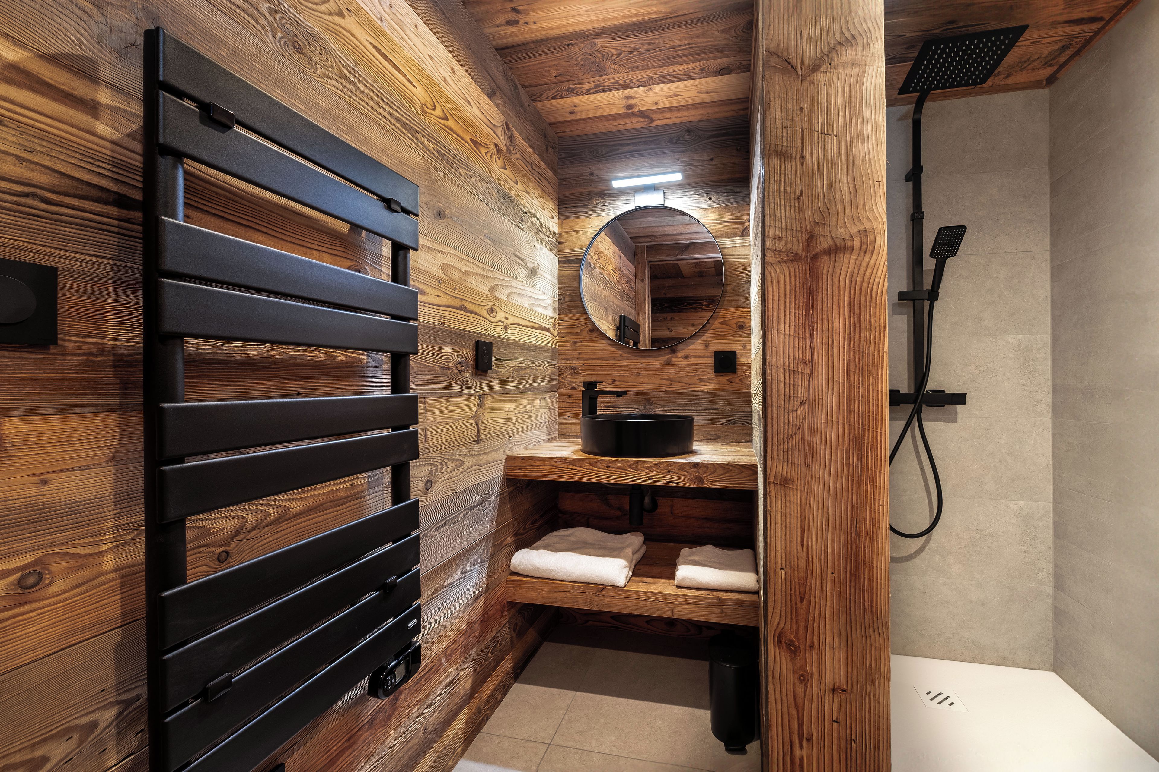 Ein Badezimmer mit Holzvertäfelung, einem schwarzen Handtuchhalter, einem runden Spiegel, einem Waschbecken und einer Dusche.