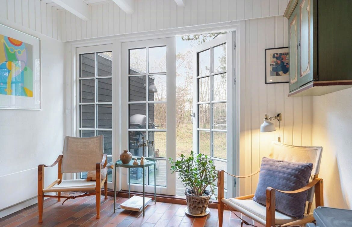 A room with a large glass door leading outside, two wooden chairs, a small table with a vase and a plant, and a lamp on the right.