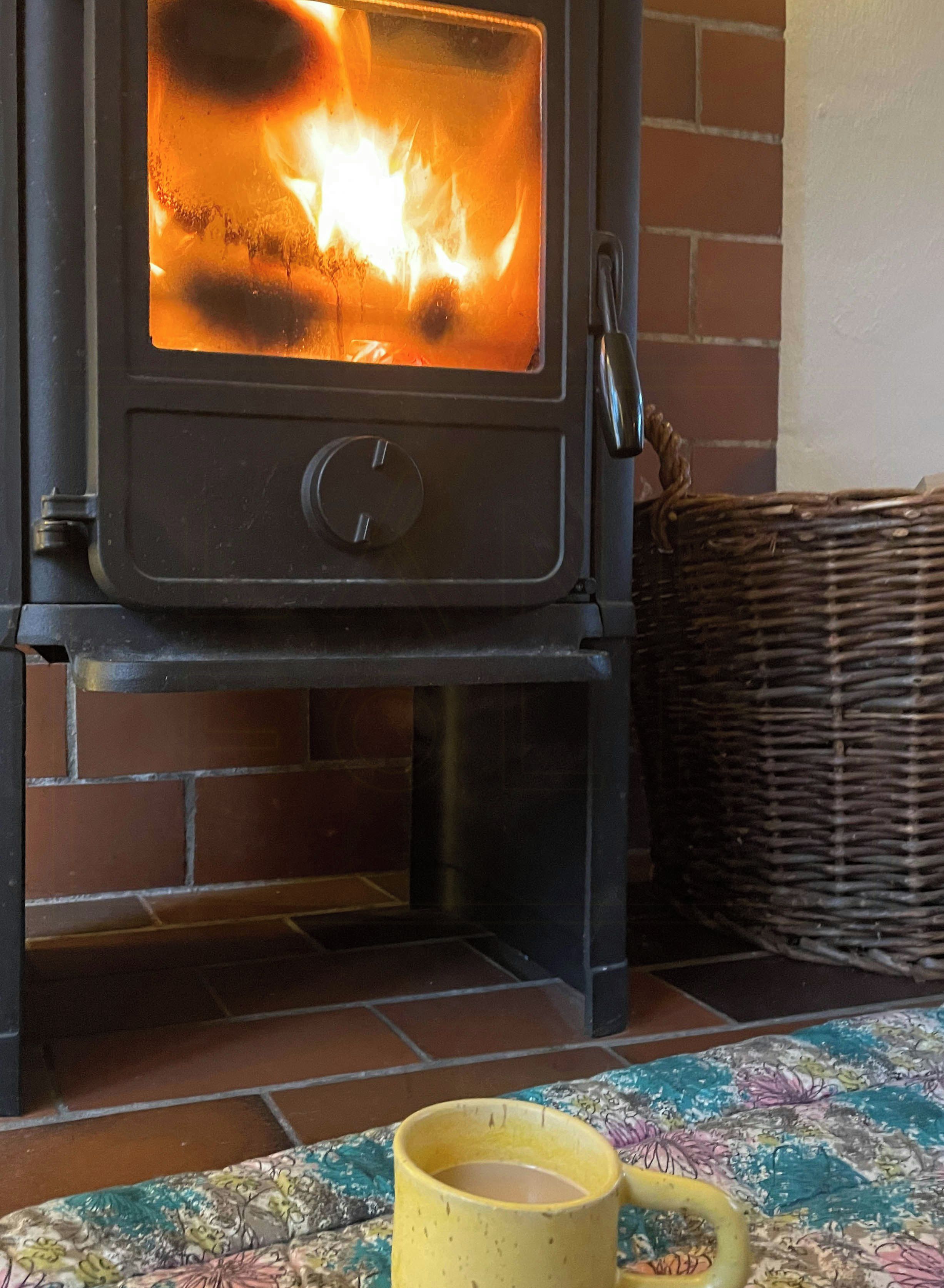 Un poêle à bois avec du feu à l’intérieur, un panier en osier et une tasse jaune posée sur une nappe à motifs.