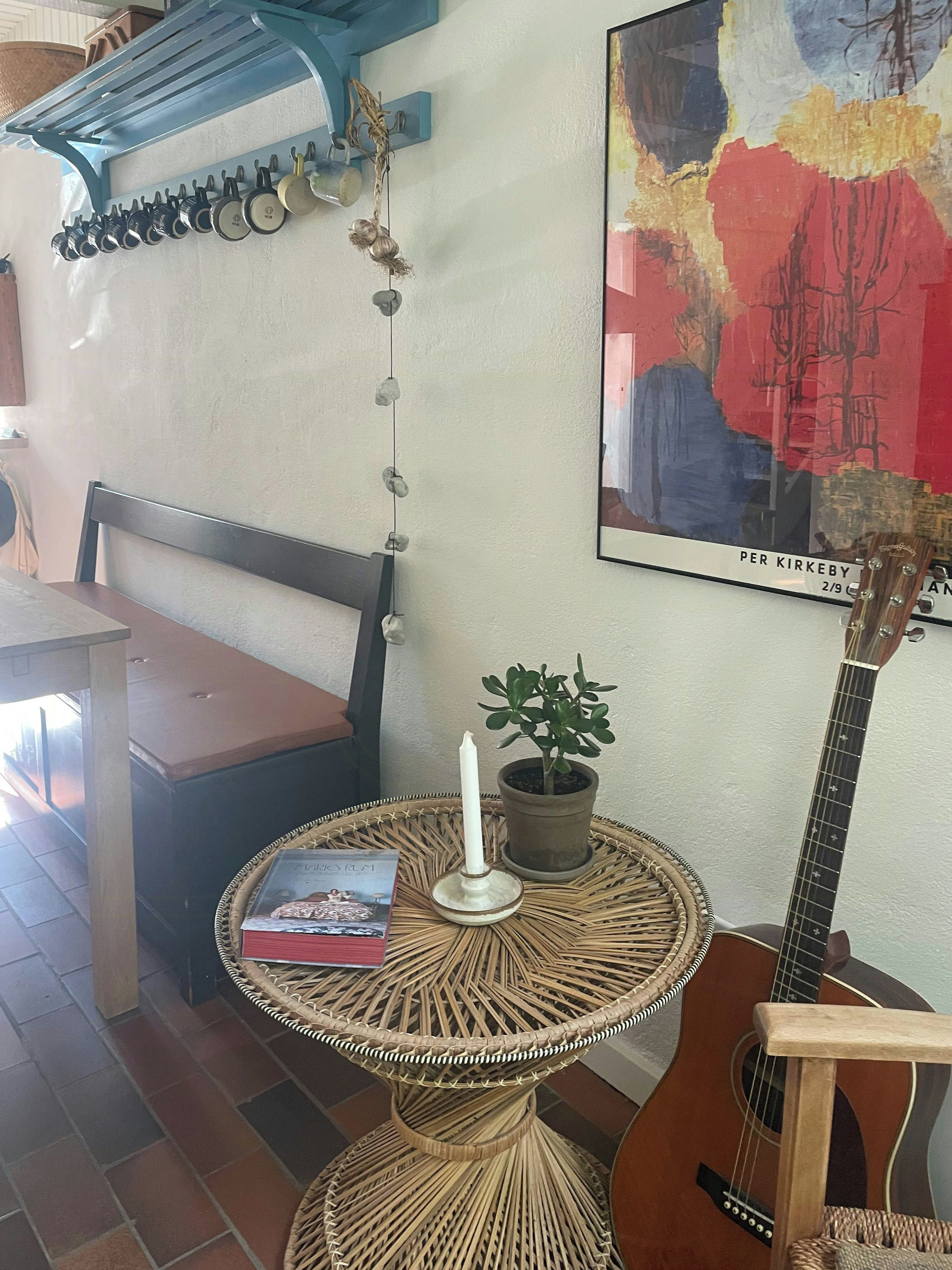 Una habitación con una guitarra, una mesa con un libro y una vela, una planta en maceta y un estante en la pared con utensilios de cocina.