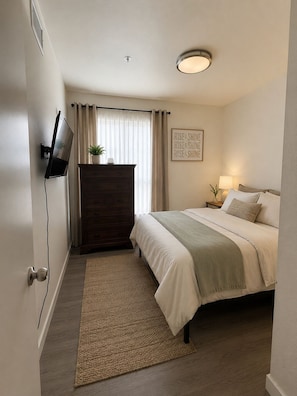 Ein Schlafzimmer mit einem Bett, einer Kommode, einem an der Wand montierten Fernseher, einer Nachttischlampe und einer Deckenleuchte.