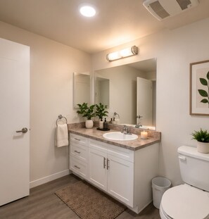 A modern bathroom with a white sink, mirror, and toilet. There are plants on the counter and a trash can beside the toilet. - Furnished 3BR Indio Apartment • 30+ Day Stays (Indio)