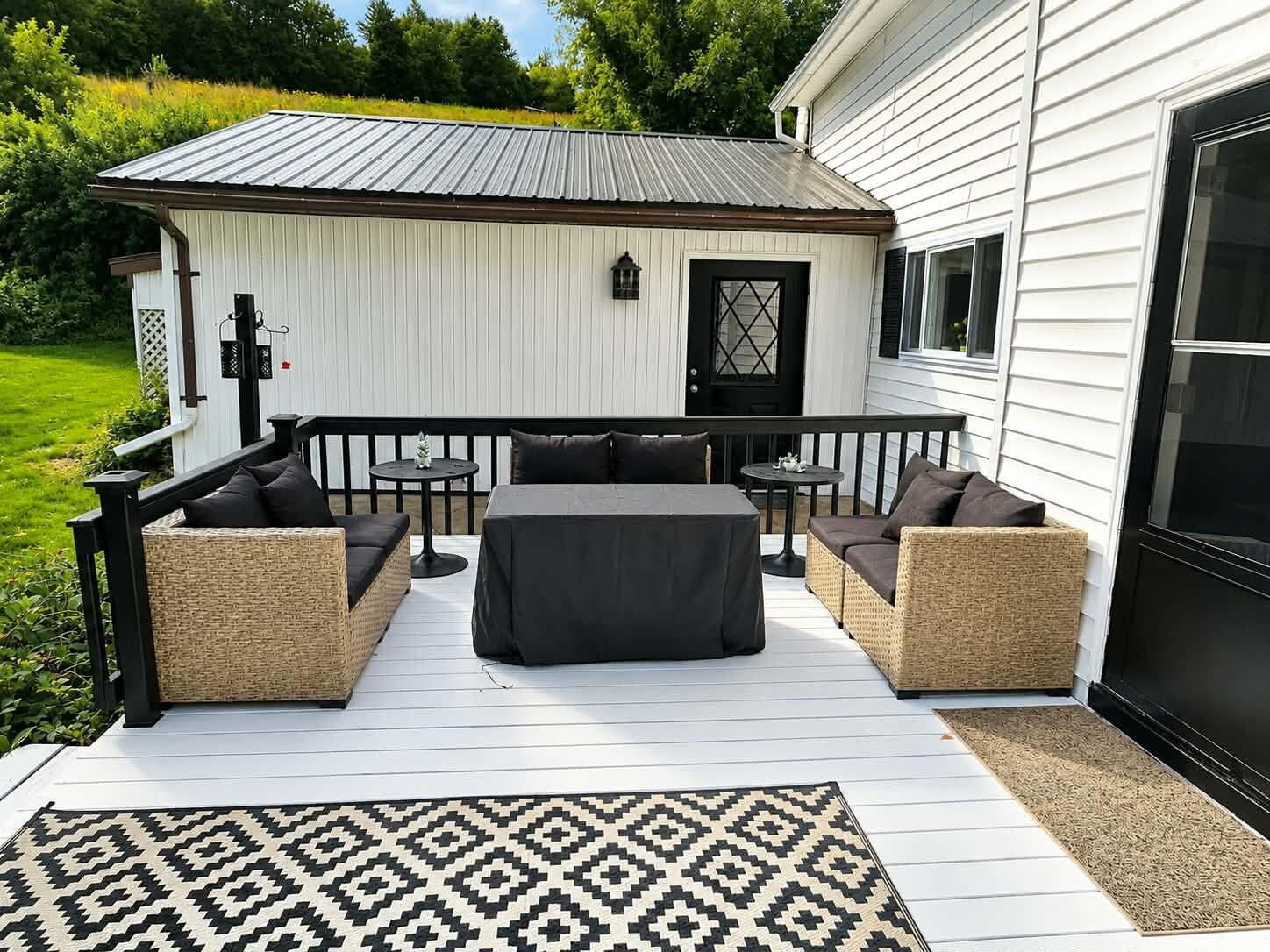 Une terrasse extérieure couverte, aménagée avec des meubles en osier et une table, un petit abri de jardin et un bâtiment blanc avec une porte noire.