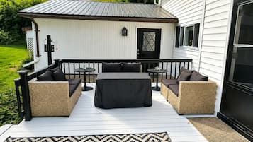 Une terrasse extérieure couverte, aménagée avec des meubles en osier et une table, un petit abri de jardin et un bâtiment blanc avec une porte noire.