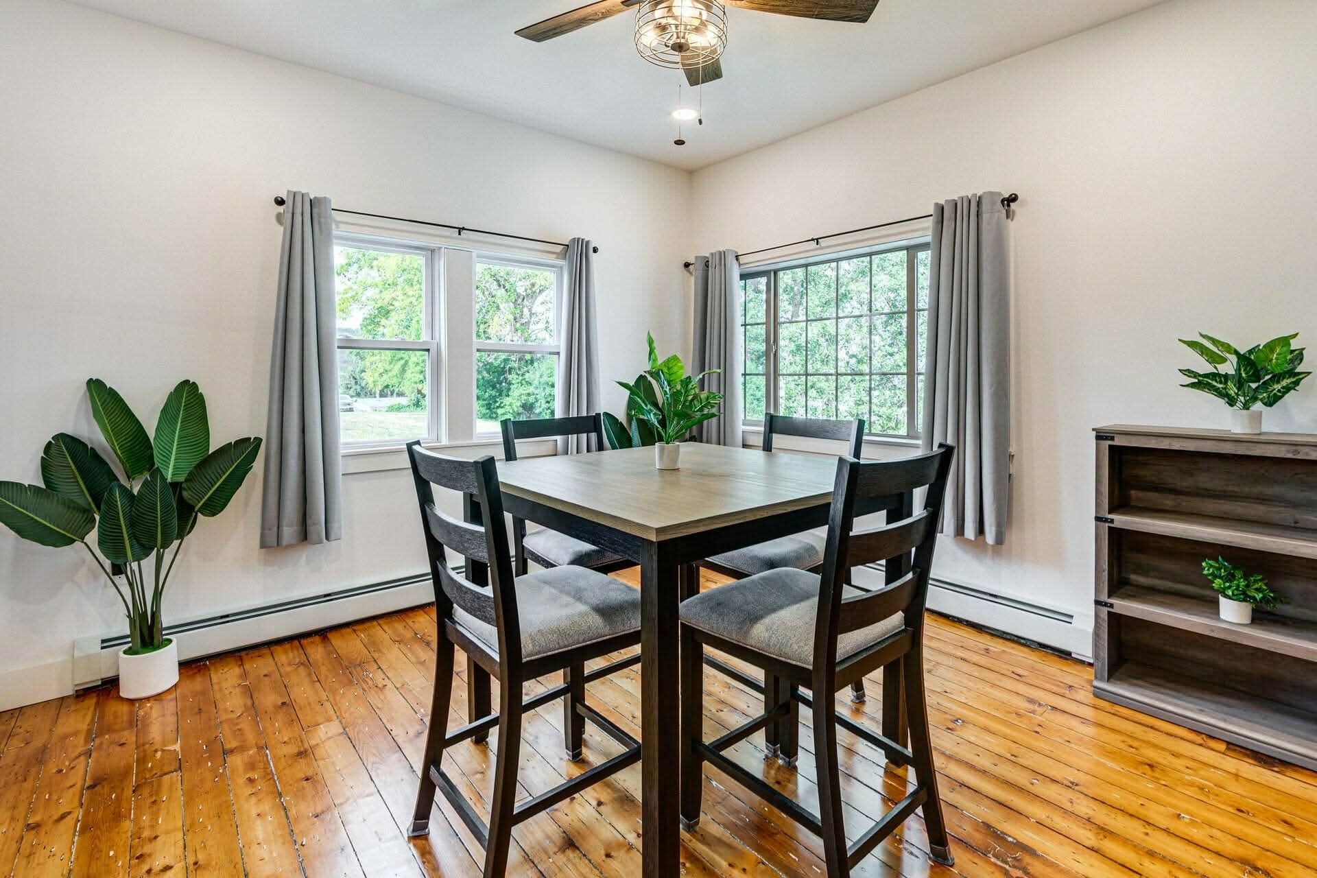 Un coin repas avec une table et des chaises en bois, de grandes fenêtres avec des rideaux et un ventilateur de plafond.