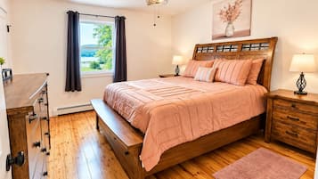 Une chambre avec un lit en bois, une table de chevet, une commode, un ventilateur de plafond, une fenêtre avec des rideaux et un tableau au mur.