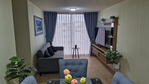 Un salon avec un canapé gris, un fauteuil bleu, une petite table ronde ornée d’une plante et un téléviseur posé sur un meuble en bois.