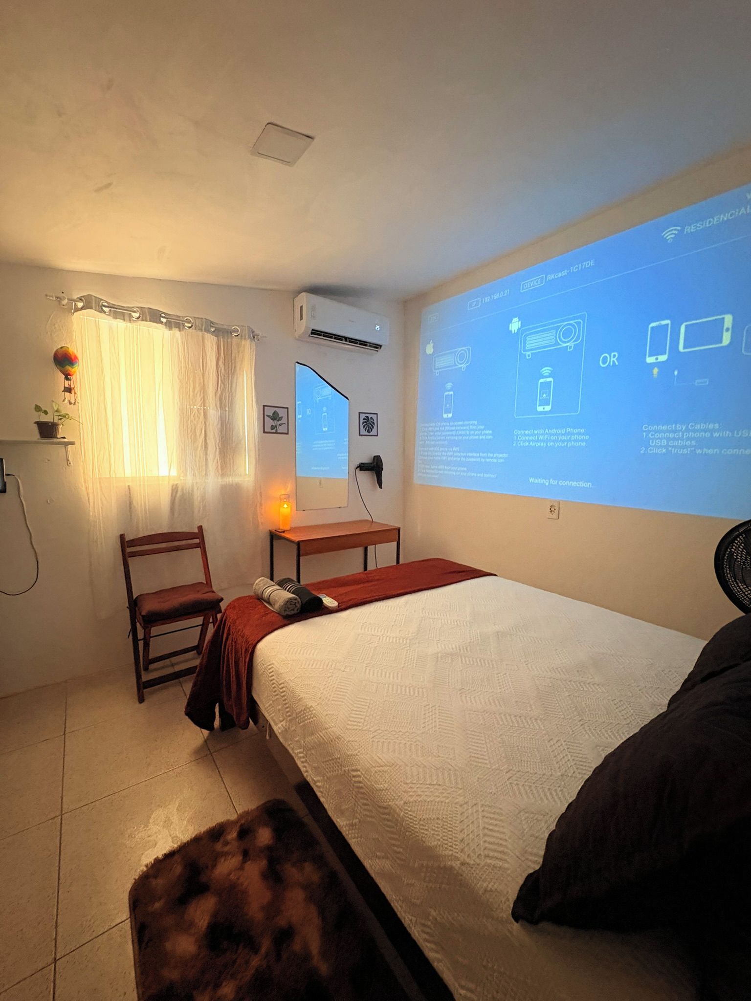 Quarto de hotel com uma cama, uma cadeira, uma escrivaninha e uma tela de projetor exibindo instruções.