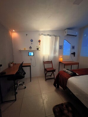 Quarto pequeno e bem iluminado, com escrivaninha, cadeira e cama. Há um monitor de computador na escrivaninha, um abajur na mesa de cabeceira e uma janela com cortinas.