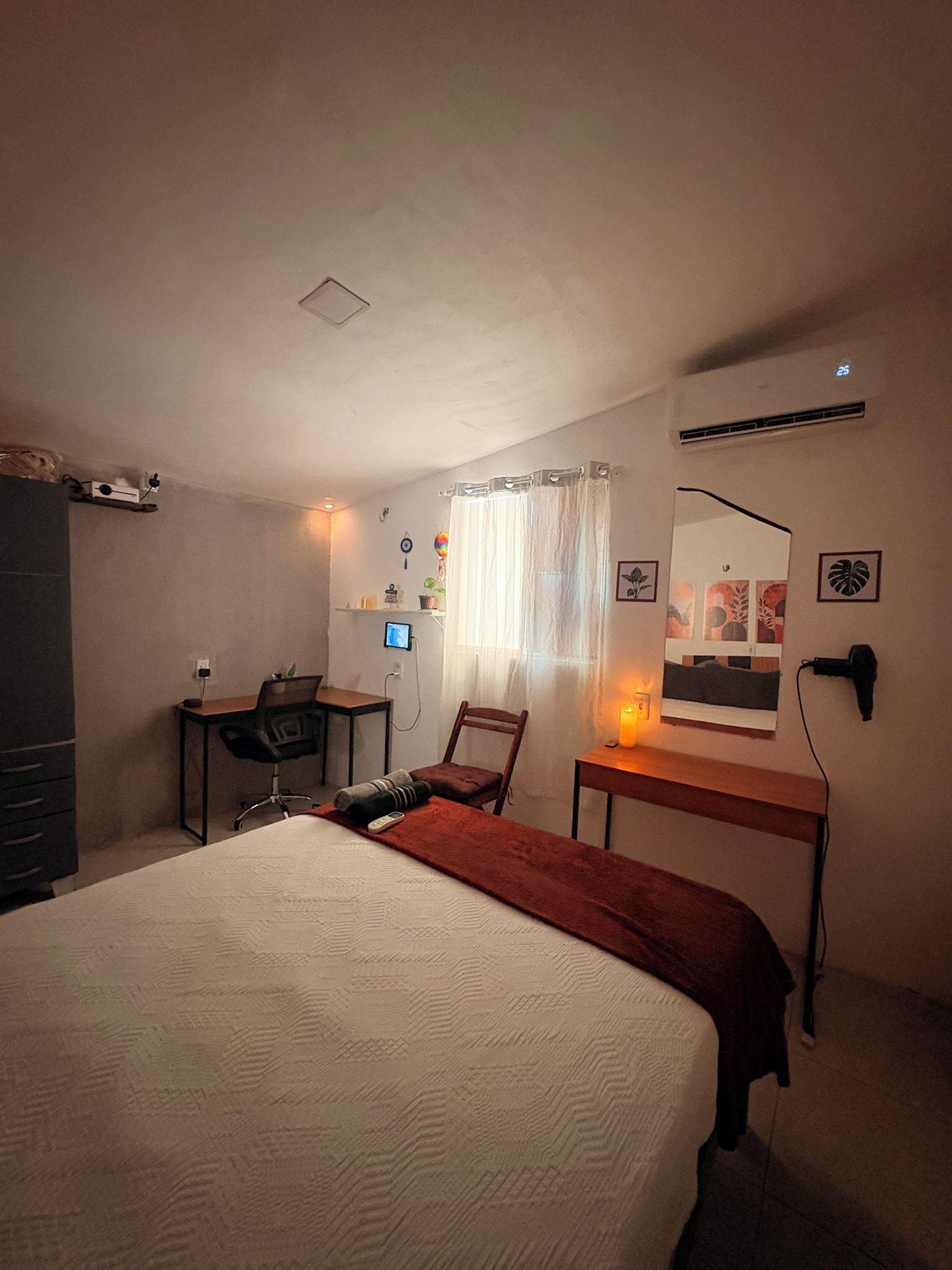 Quarto com cama, escrivaninha, cadeira, mesa de cabeceira com abajur e ar-condicionado fixado na parede.