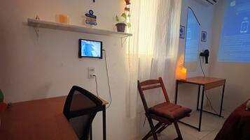 Quarto com escrivaninha, cadeira e uma mesinha. Há uma televisão na parede, um vaso de plantas e uma janela com cortinas.