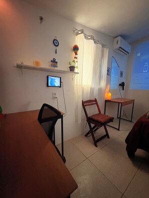Quarto com escrivaninha, cadeira e uma mesinha. Há uma televisão na parede, um vaso de plantas e uma janela com cortinas.