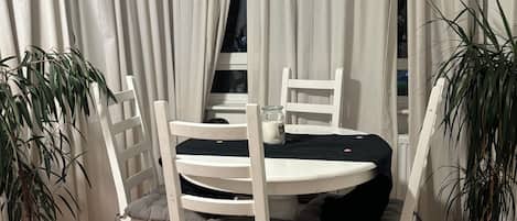 A dining area with a round table and white chairs, surrounded by white curtains, with potted plants on either side.