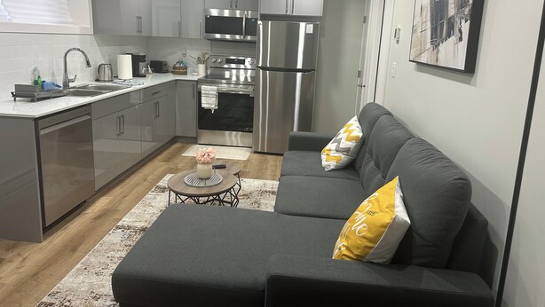 A modern kitchen with stainless steel appliances, a grey sofa with cushions, and a small round table with a vase. - Bethel Place - brand new suite in Evergreen! (Saskatoon)