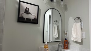 A bathroom with a white sink, a framed picture of a bear in a bathtub, a mirror, and three light fixtures above the mirror. - Bethel Place - brand new suite in Evergreen! (Saskatoon)