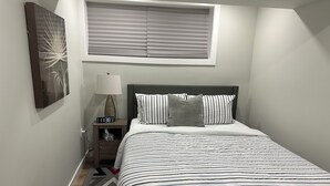 A neatly made bed with striped bedding, a bedside table with a lamp, a wall clock, and a window with blinds. - Bethel Place - brand new suite in Evergreen! (Saskatoon)