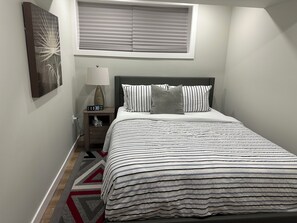 Een net opgemaakt bed met gestreepte lakens, een nachtkastje met een lamp, een wandklok en een raam met zonwering.