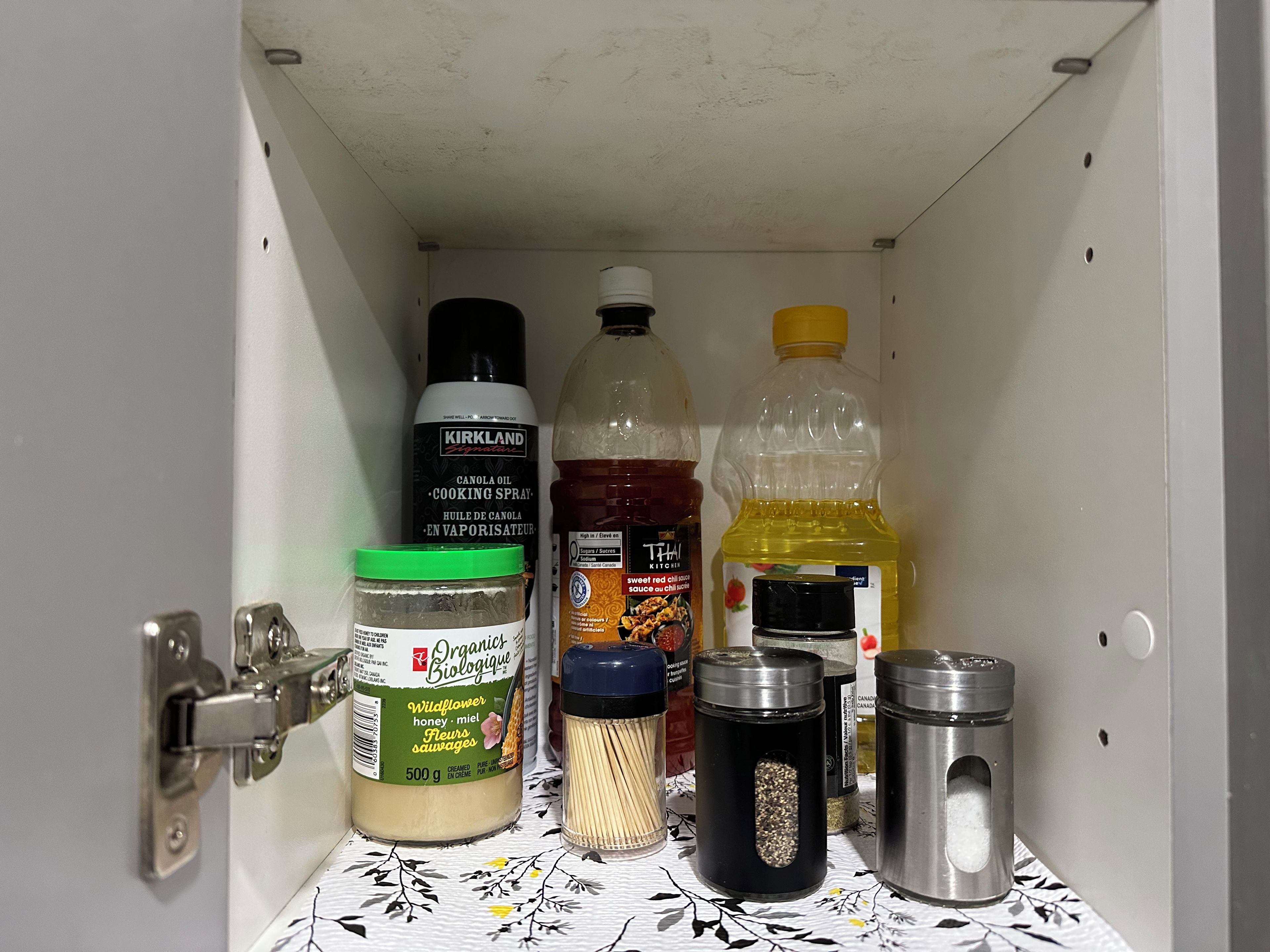A kitchen cabinet with various food items including a canola oil spray, a jar of honey, a bottle of Thai sauce, and a container of sugar.