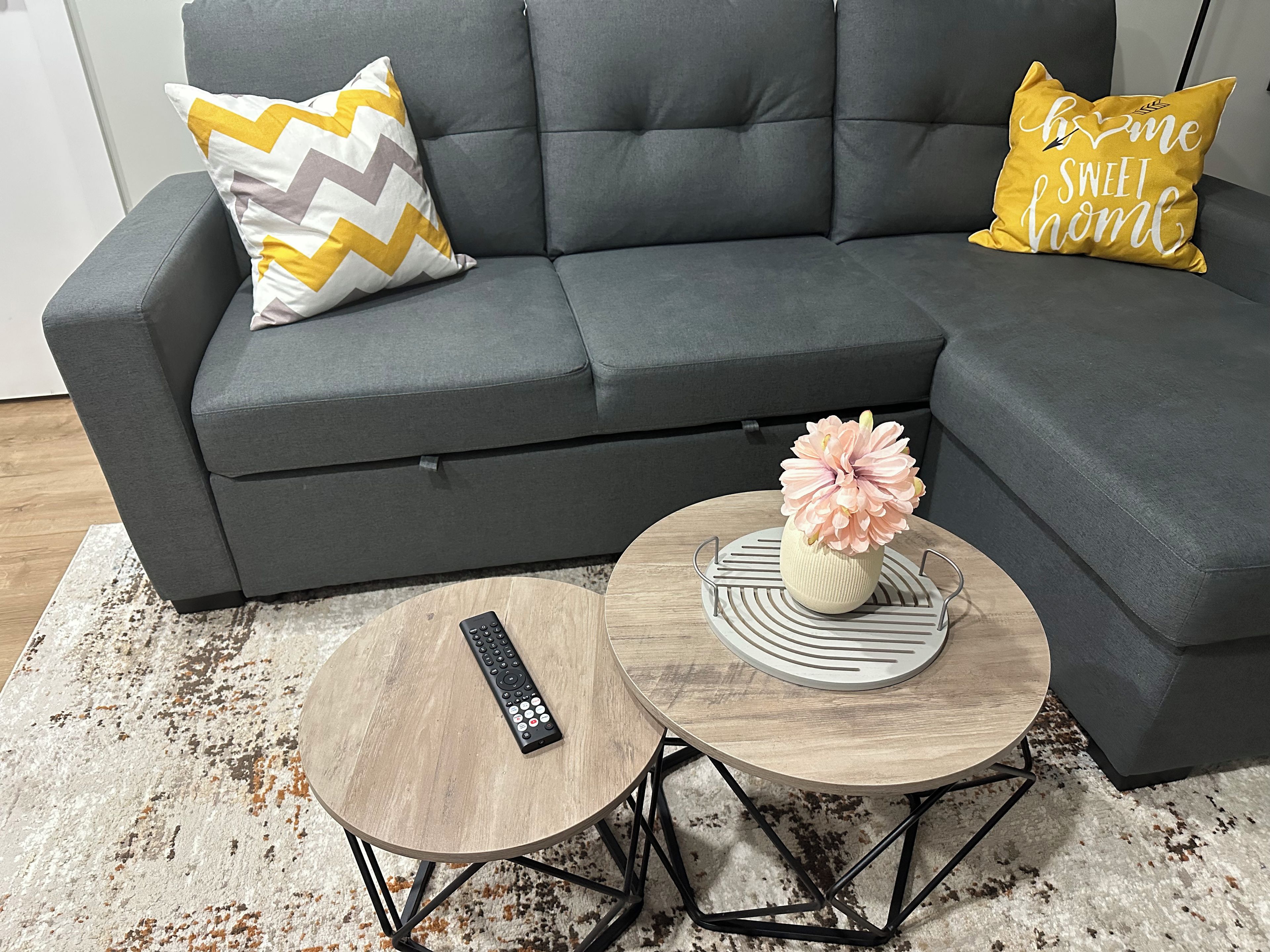 A grey sofa with two pillows, a remote control on a small round table, and a vase with flowers on a larger round table.