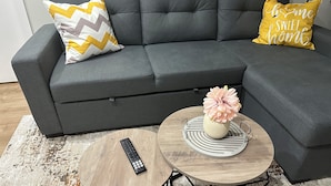 A grey sofa with two pillows, a remote control on a small round table, and a vase with flowers on a larger round table. - Bethel Place - brand new suite in Evergreen! (Saskatoon)