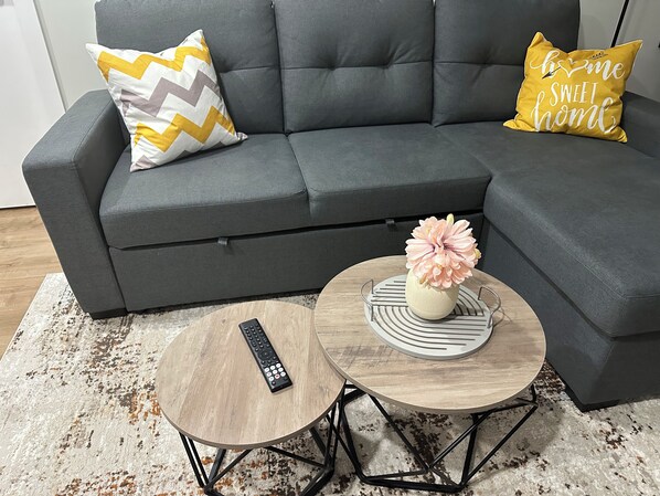 Un canapé gris avec deux coussins, une télécommande posée sur une petite table ronde et un vase rempli de fleurs sur une table ronde plus grande.